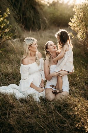 Three people in white dresses share a candid moment together outdoors during golden hour in a grassy field.