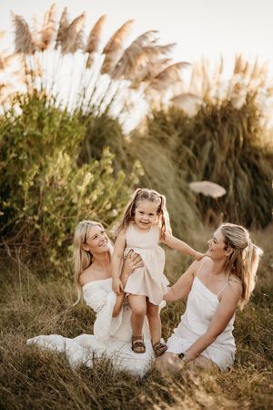 Three people in white dresses sit together in a grassy field with pampas grass in the background at sunset.