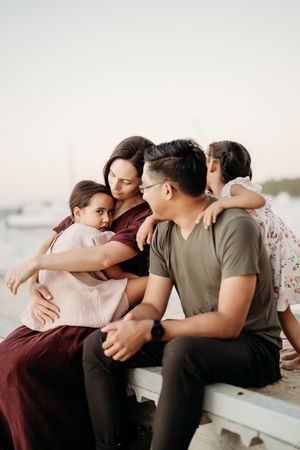 Family sits together in tender embrace on dock at sunset with water visible in background.