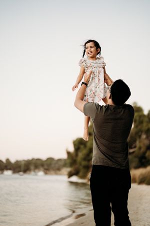 A parent lifts a small child wearing a floral dress by a lakeside at sunset while sharing a joyful moment together.