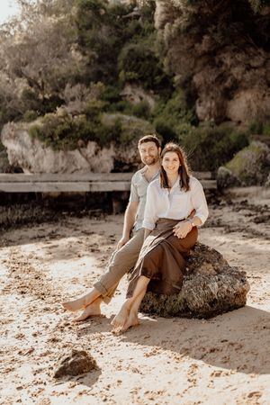 A couple lounges together on a sandy beach near rocky cliffs during a romantic seaside photoshoot.