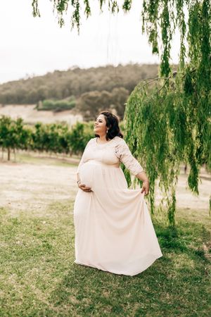 Elegant maternity photo at a vineyard with flowing cream lace dress under a drooping willow tree.
