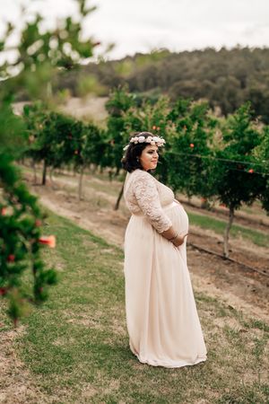 Expectant mother wearing floral crown and cream lace dress poses in rustic vineyard setting during maternity photoshoot.