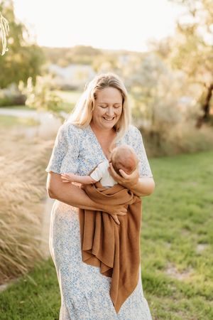 A parent in a floral dress holds a newborn baby wrapped in a brown blanket during golden hour in a grassy field.