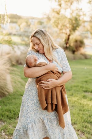 A tender moment between mother and newborn baby outdoors in a sunlit garden with pampas grass and greenery.