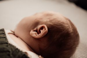 Close-up view of a baby's ear and head resting on a soft surface near a green sweater sleeve.