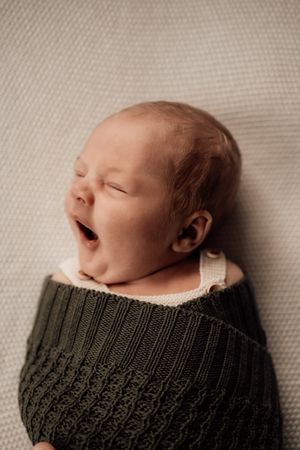 A sleeping newborn baby swaddled in a dark green knit blanket against a neutral cream background.