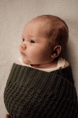 Baby wrapped in dark green knit blanket lying on textured beige surface in soft portrait lighting.
