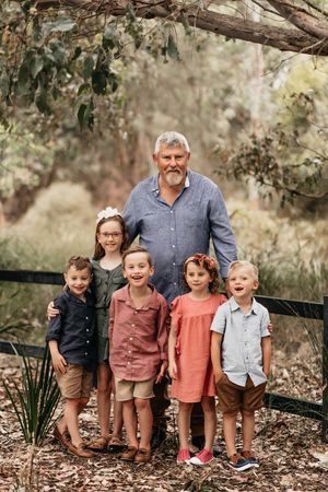 A group photo outdoors in a wooded setting with adults and children standing together under leafy branches.