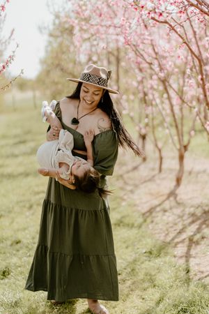 Family photoshoot among blooming cherry blossom trees with flowing olive dress and boho hat in springtime.