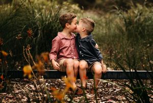 Two young children share a sweet moment while sitting together on a bench surrounded by wildflowers in a garden setting.