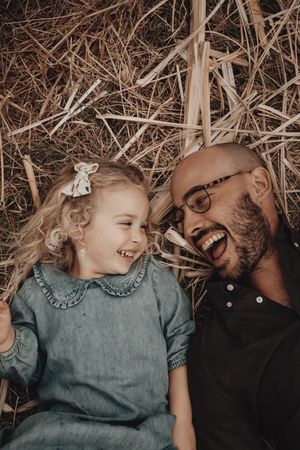 A joyful father and daughter share laughter while lying on hay together in a heartwarming candid moment.