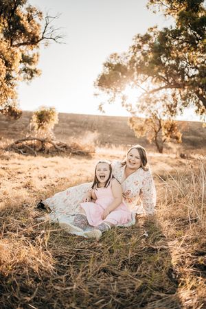 Two people in feminine outfits sit together on a blanket in a sun-drenched field during golden hour at sunset.