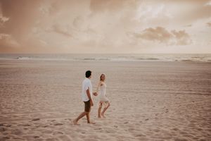 A couple walks hand in hand on a sandy beach during sunset with warm sepia tones in the sky.