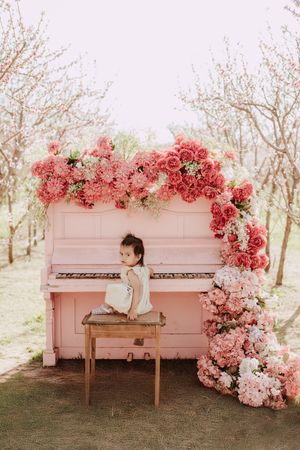 A vintage pink piano decorated with cascading pink and coral flowers in a dreamy outdoor garden setting.