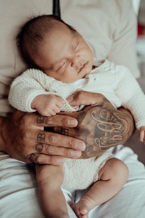 A sleeping newborn in a white sweater holds onto tattooed hands in a tender moment.