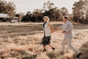 Two people holding hands and dancing together in a golden field at sunset with rustic buildings in the background.