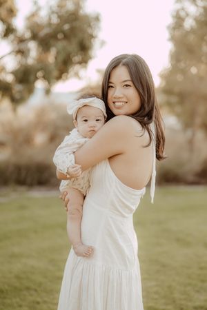A parent and child wearing white dresses embrace while standing on a grassy field during golden hour.