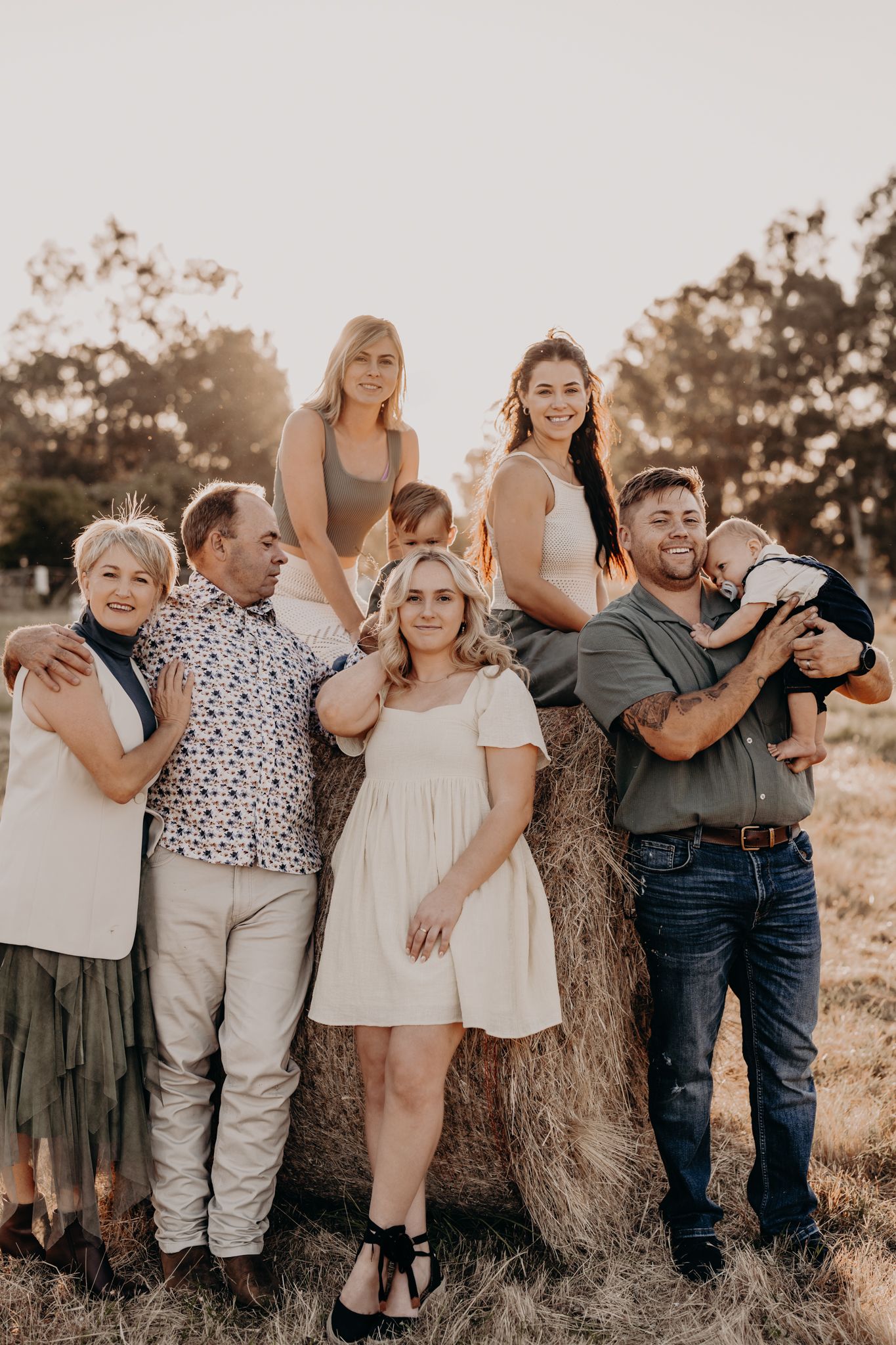 A family poses together outdoors at sunset against a rustic hay bale backdrop in casual coordinating outfits.