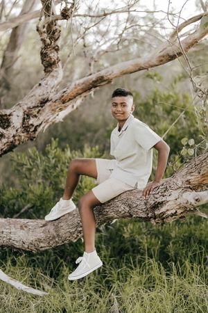 Someone wearing a cream colored outfit sits casually on a fallen tree branch in a natural outdoor setting.