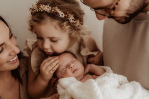 Intimate family moment as toddler with floral crown gently touches sleeping newborn swaddled in white blanket.