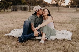 A couple sitting on a white blanket in a grassy field during golden hour shares an intimate moment together.