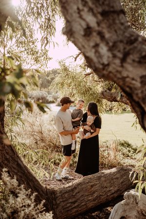 A family shares an intimate moment together framed by a large tree branch in a sunlit outdoor setting at dusk.