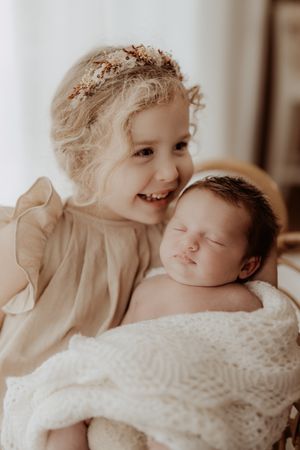 A child wearing a floral crown smiles while holding a sleeping newborn baby wrapped in a white knit blanket.