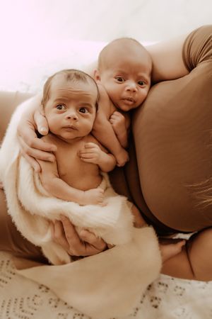 Two infant babies cuddle together on a bed, one wrapped in a soft white blanket, in a warm sepia-toned photograph.