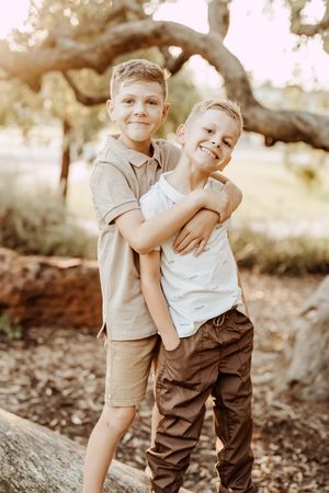 Two young boys in casual clothing stand together outdoors smiling and hugging in warm sunlight beneath a tree.