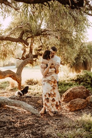 Romantic couple embrace under an olive tree as golden sunlight filters through branches during an outdoor photoshoot.
