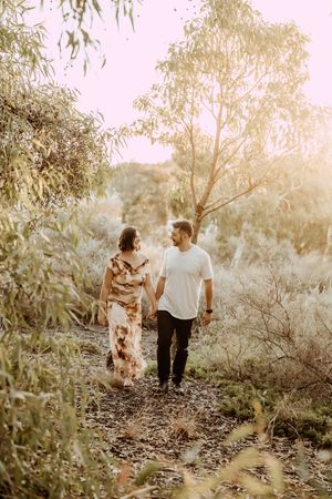A couple walks hand in hand along a nature trail during golden hour with sunlit trees and foliage surrounding them.