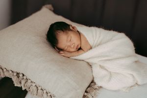 A sleeping newborn wrapped in white cloth resting peacefully on a textured white pillow.