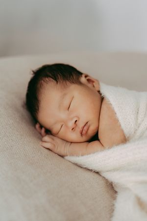 Sleeping newborn wrapped in soft white blanket rests peacefully on textured fabric.