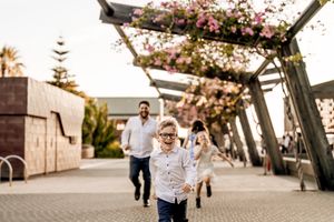 Child runs joyfully under a wooden pergola covered in pink flowering vines as someone follows behind at sunset.