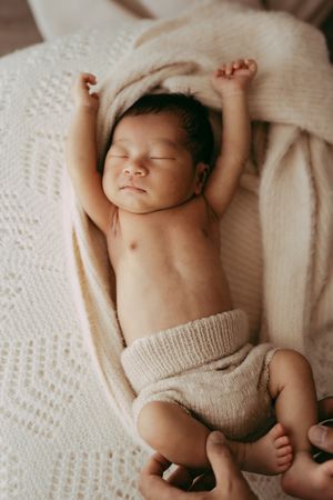 Peaceful sleeping newborn stretches arms while lying on white knitted blanket wearing beige shorts.