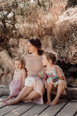Three figures in soft pastel dresses sit together on a wooden deck against a rocky natural backdrop in warm lighting.