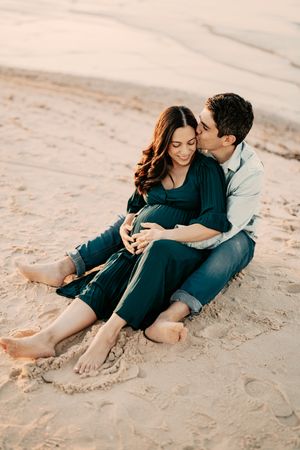 A couple sits together intimately on a sandy beach during a romantic sunset photo session.