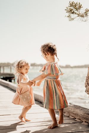 Two children in summer dresses dance barefoot together on a wooden dock by the water on a bright sunny day.