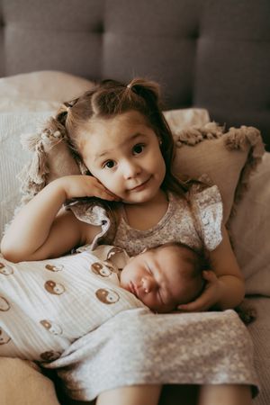 Two children in matching gray outfits cuddle together on a couch with patterned blanket and pillows.