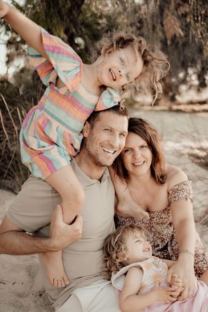 A family sits together outdoors smiling and cuddling on a sunny day wearing pastel and neutral tones.