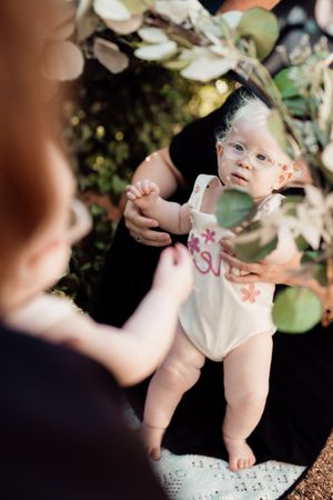 A baby in a white dress sits among greenery in a garden photoshoot setting.