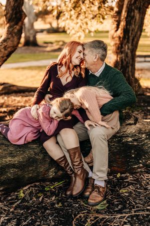 A family shares a tender moment sitting together on a fallen tree trunk during a golden autumn afternoon.