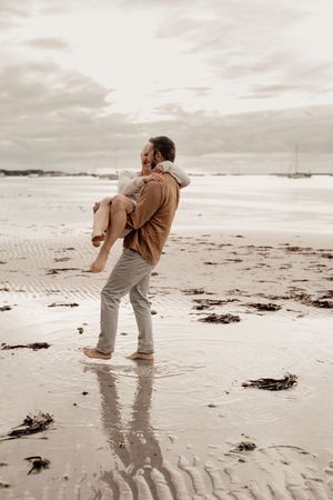 A couple in casual attire embraces while standing on a misty beach during golden hour, their reflection visible in the wet sand.