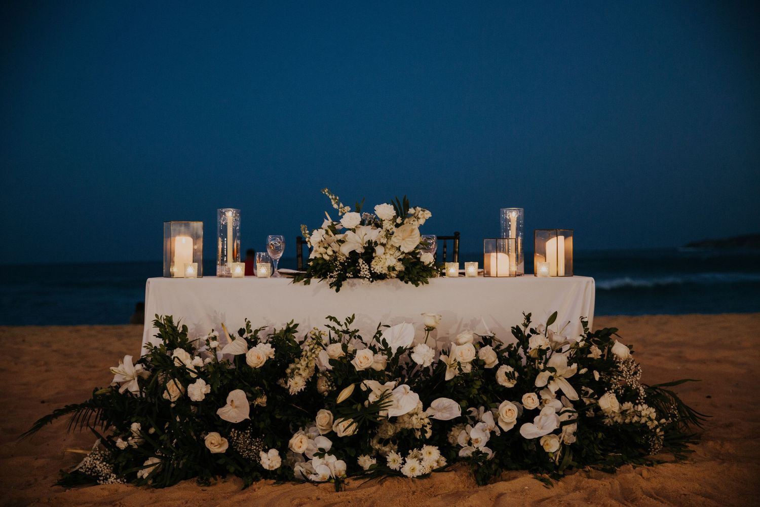 White floral arrangements decorate a beachside wedding ceremony setup against a blue sky.