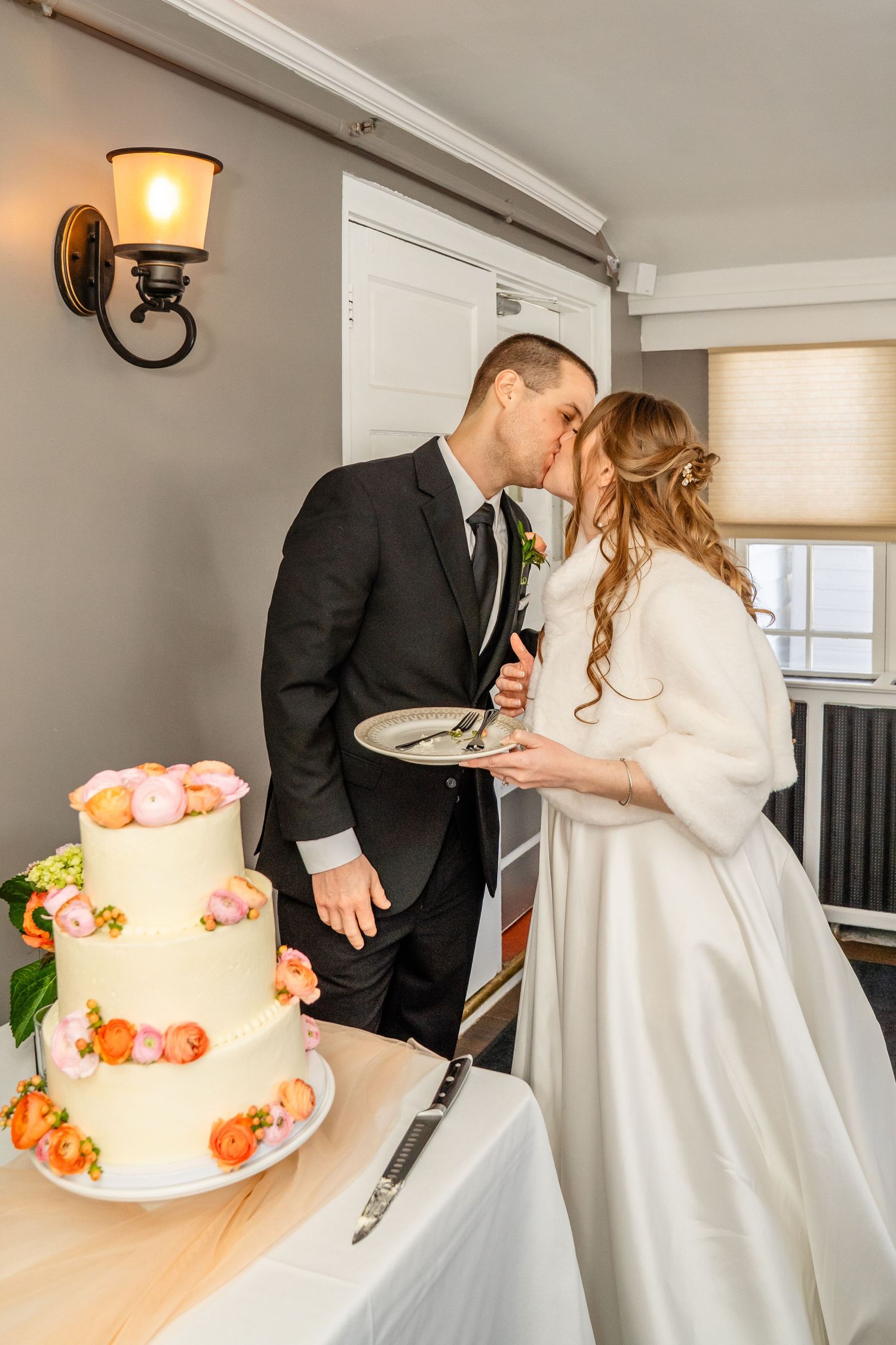 A couple cuts their white wedding cake decorated with peach roses flowing down the side during their wedding reception.