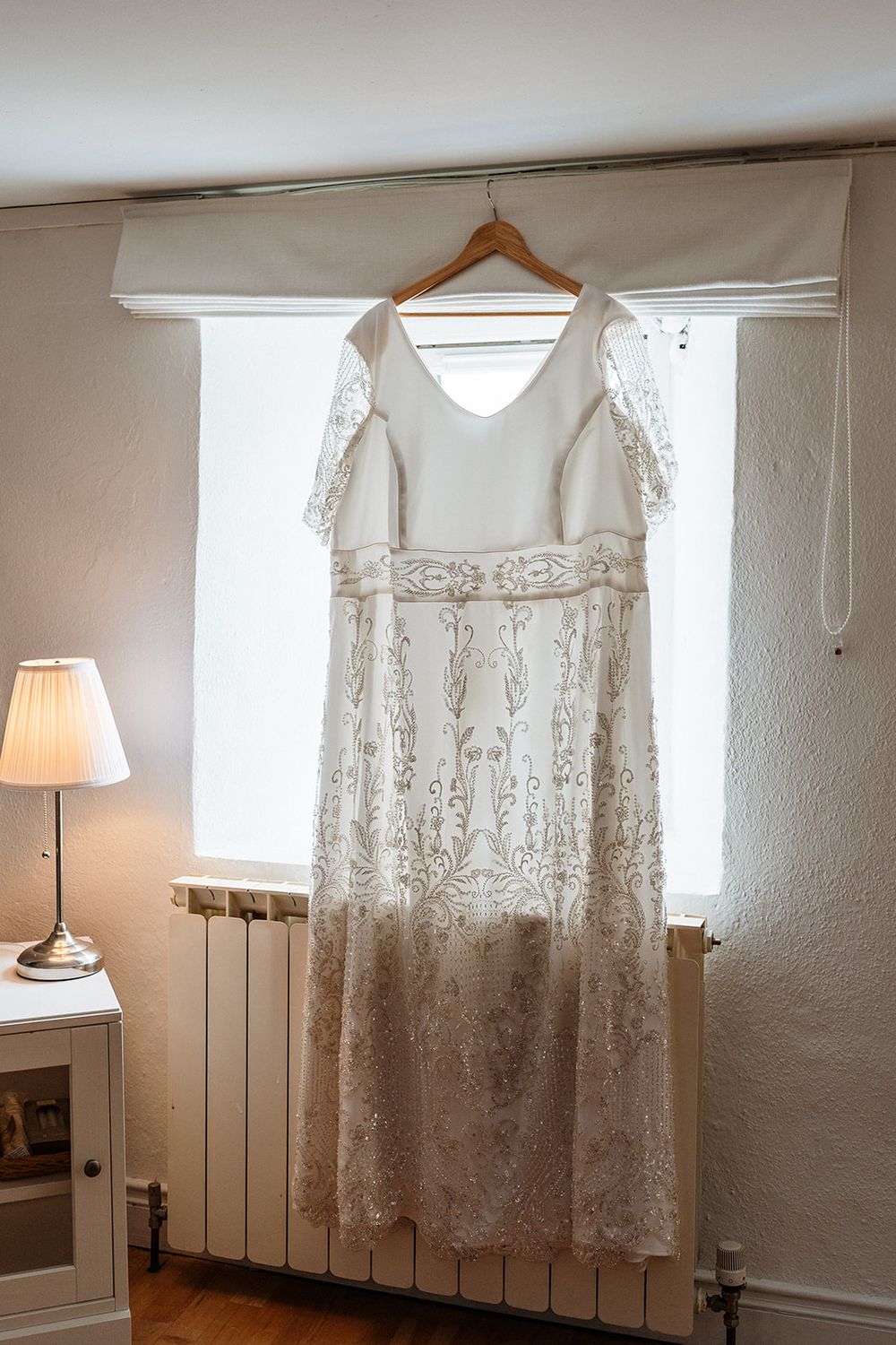 A vintage-style white lace wedding dress with intricate embroidery hangs on a wooden hanger near a window and table lamp.