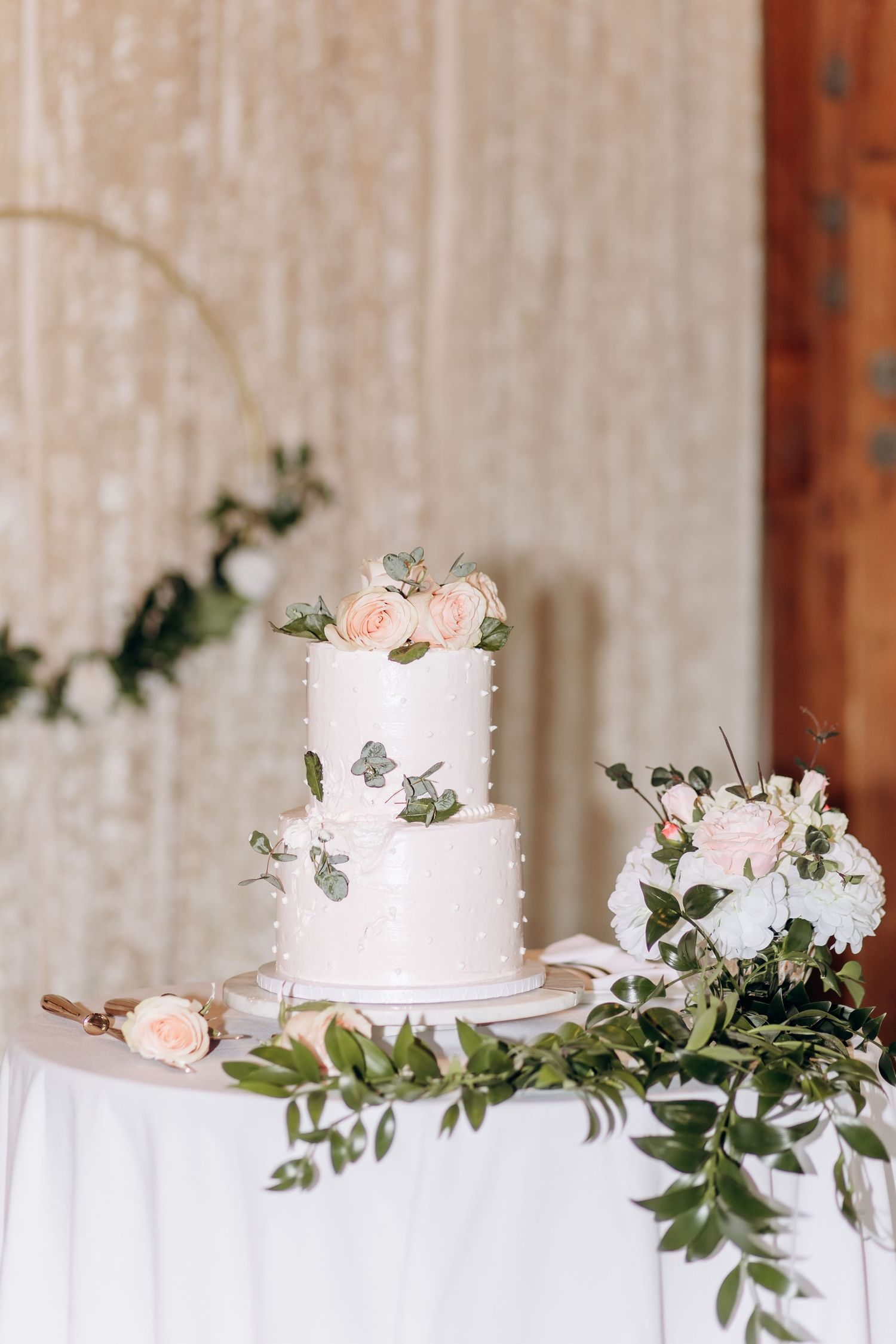 Elegant white wedding cakes decorated with pink roses and greenery displayed on a table with draped fabric.