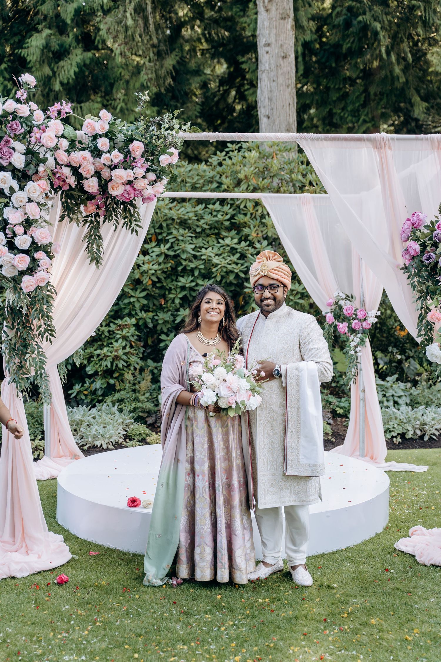 Outdoor wedding ceremony setup with white drapes, floral arrangements, and guests in traditional attire.