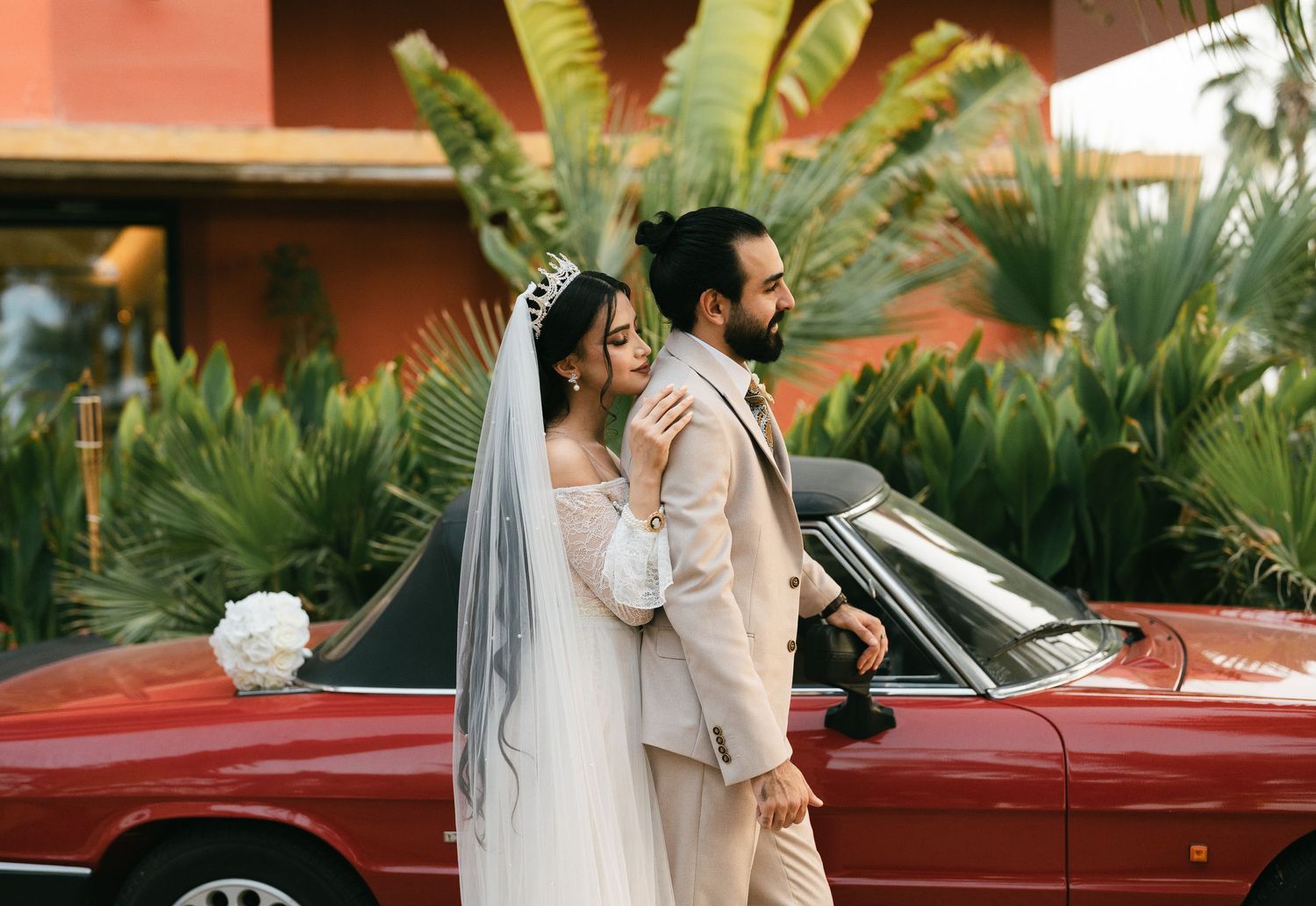 A happy couple in wedding attire stands next to a red vintage convertible car with tropical plants in the background.